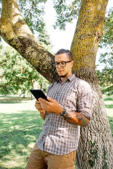 Handsome young man standing near the tree and using digital tablet in summer park.