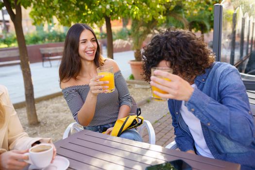 Couple of friends toasting with orange juices while having a drink with their multi-ethnic group of friends at the outdoor table of a bar.