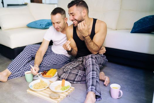 Gay couple eating together sitting on their living room floor. Homosexual lifestyle concept.