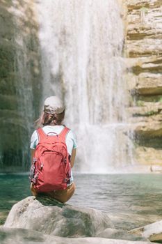 Backpacker young woman sitting on stone and enjoying view of waterfall on sunny summer day.