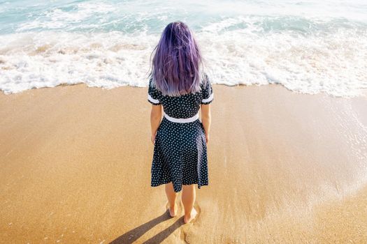 Young woman with violet hair standing on sand beach and enjoying view of sea, rear view.