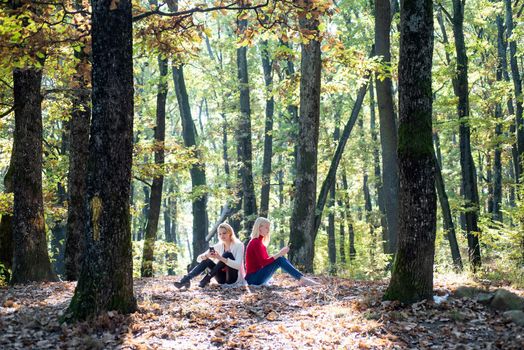 Portrait of two active young girls holding mobile phone and sitting on fallen leaves outdoor. Young women in Autumn. Beautiful fall time in nature