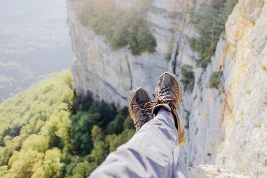 Explorer young man sitting on edge of cliff over valley, view of legs, pov.