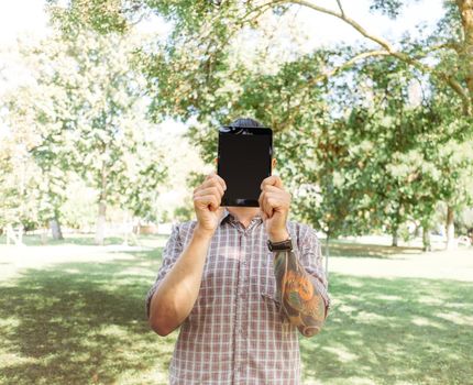 Young man holding digital tablet with empty screen in front of his face in summer park.