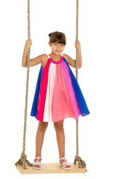 Cute happy girl standing on wooden swing. Full length shot of lovely girl swinging against white background. Beautiful child in colorful summer dress and sandals having fun outdoor