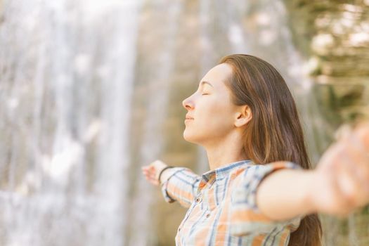 Young woman with closed eyes relaxing with raised arms on background of waterfall in summer outdoor, side view.