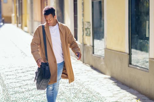 Young black man walking down the street carrying a briefcase and a smartphone. Cuban guy in urban background.