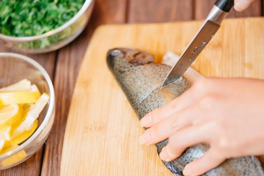 Unrecognizable woman cutting raw fish trout on wooden board for dish in the kitchen, view of hands close-up.