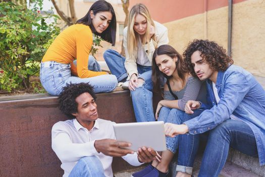 Multi-ethnic group of students looking at something on a digital tablet sitting outdoors.