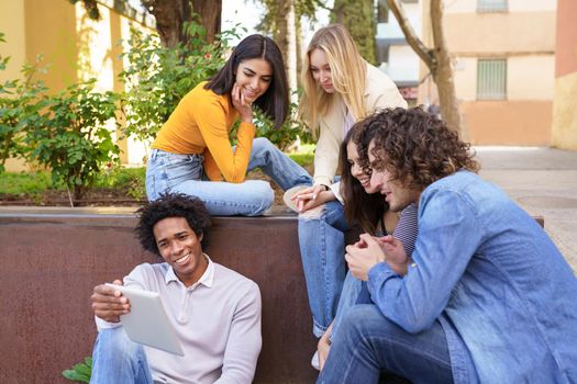 Young black man showing something on his digital tablet to his group of friends sitting on some steps in the street.