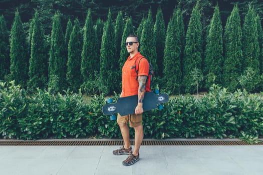 Handsome tattooed guy in sunglasses standing with longboard in summer park.