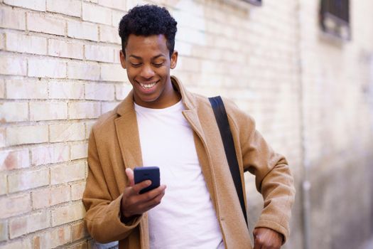 Young black man typing on his smartphone leaning against a brick wall in the street. Cuban man on urban background.