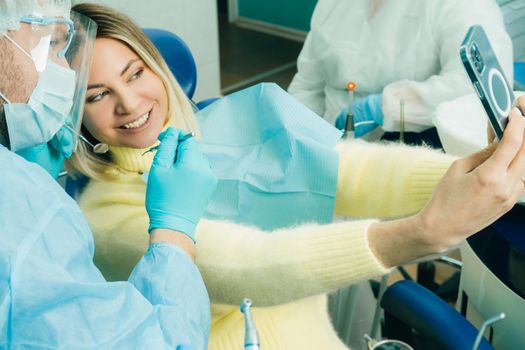 the dentist in a protective mask sits next to the patient and takes a photo after work.