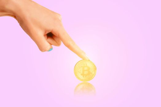Female hand pointing to glowing gold coin bitcoin on a pink background.