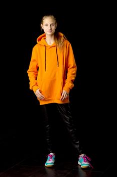 Full length shot of stylish teenage girl. Joyful girl with ponytail wearing oversized orange hoodie and black leather pants standing against black background in studio