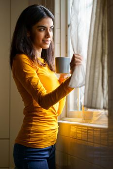 Persian woman drinking coffee at home while looking through the window