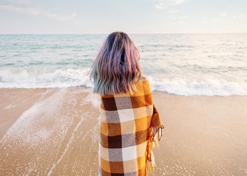 Young woman wrapped in a plaid standing on sand beach and looking at sea, rear view. Summer vacations.