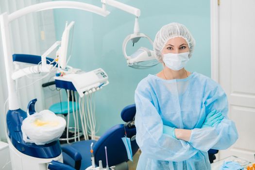 a female dentist wearing a medical mask and rubber gloves poses for the camera and folds her arms in her office.