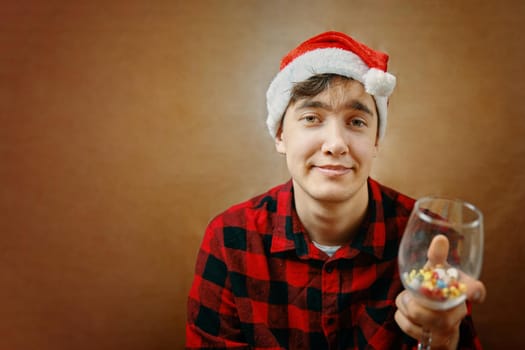 Guy in Santa Claus hat holds a glass with various pills and drugs. Fighting the coronavirus on holiday. Holidays spending under quarantine. Man with grimace on face. New year mood.