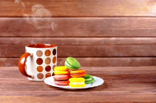 Sweet colorful macarons dessert on a plate and cup of hot beverage on wooden background.
