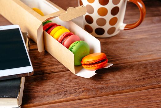 Sweet macarons and cup of drink on a wooden table near the smartphone and notepad.