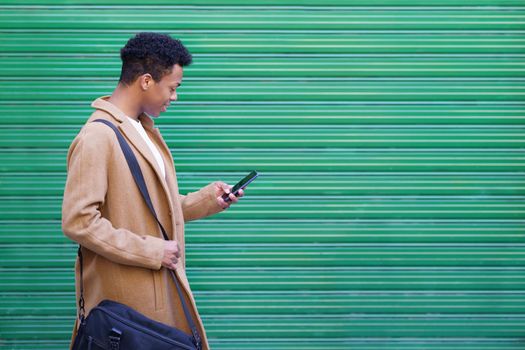 Black young man looking at his smartphone near green blinds. Cuban guy smiling in urban background.