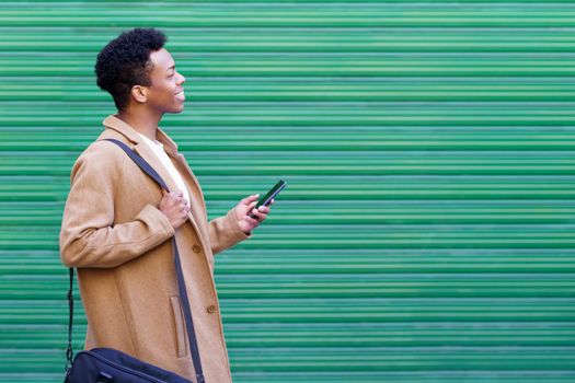 Cuban young man looking forward using his smartphone near green blinds. Black guy smiling in urban background.