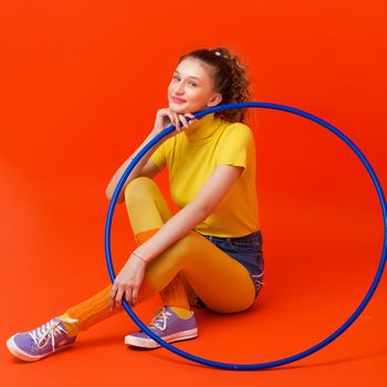 Happy fitness girl doing exercises with hula hoop. Portrait of cheerful girl sitting on floor against red background. Student looking stylish in turtleneck blouse, leggins, shorts and socks gaiters
