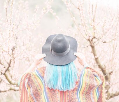 Young woman with blue hair in hat walking in blossoming cherry tree garden in spring, rear view.