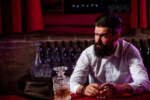 Handsome pensive man is holding a glass of whiskey and looking away while sitting in bar indoors