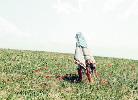 Stylish young woman with blue hair wearing in poncho walking on meadow of tulips.