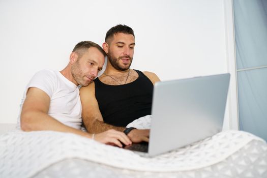 Gay couple consulting something on their laptop while lying on the bed. Homosexual Lifestyle concept.