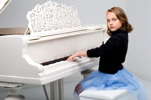 Beautiful teenage girl playing grand piano. Pretty blonde girl in stylish clothes learning to play music instrument. Teenage child looking at camera against gray background