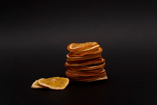 Closeup at stack of sliced dried dehydrated oranges and two sliced of dried oranges nearby, at black background, copy space, no people.