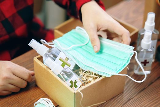 Men's hands put medical masks in a gift box with alcohol antiseptics. Sticker with a red cross on the sanitizers. Gift in quarantine on a wooden table.