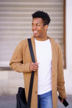 Young black man walking down the street carrying a briefcase and a smartphone in his hand. Cuban guy smiling in urban background.