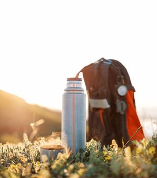 Thermos and cup of hot beverage near the backpack on green grass outdoor in summer.