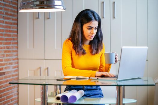 Female architect working at home with a laptop and blueprints. Persian woman. Persian woman with a coffee mug.