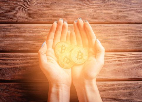Many glowing gold bitcoins in female hands on a wooden background, top view.