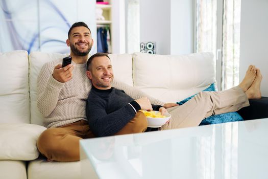 Gay couple sitting on the couch at home watching something on TV and having a snack. Homosexual relationship concept.