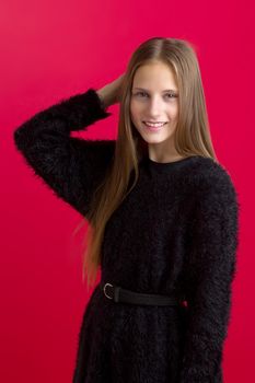 Beautiful teenage girl touching her long hair. Portrait of charming smiling teenager wearing black stylish oversized sweater with leather belt standing on red background