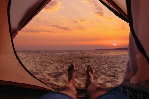 Young man resting in a tent on sand beach near the sea at sunset, point of view.