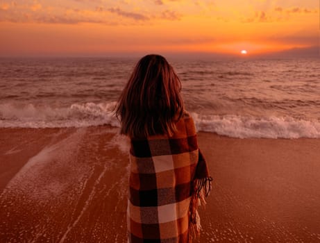Young woman wrapped in a plaid standing on sand beach and looking at sea at sunset. Summer vacations.