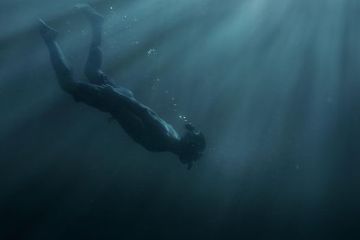 Young man freediver swimming with mask and snorkel underwater in deep sea among sunbeams.