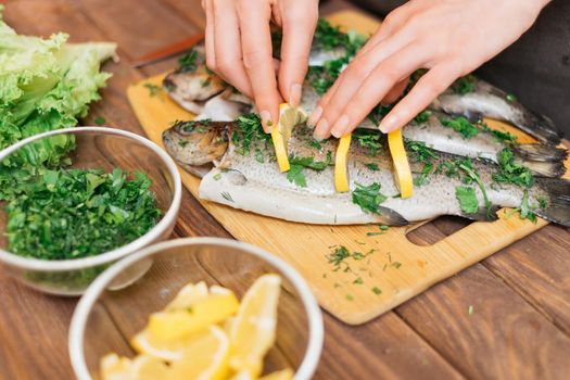 Woman preparing fish trout with lemons and greenery on wooden table, view of hands.