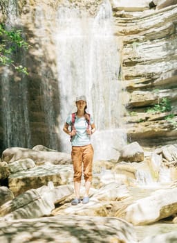 Traveler young woman with backpack standing on background of waterfall and looking at camera on sunny summer day.