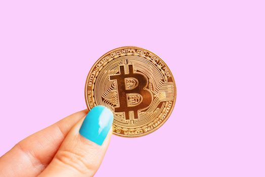 Female hand holding gold coin bitcoin on a pink background, symbol of crypto currency.