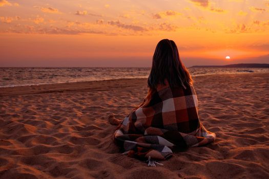 Unrecognizable woman wrapped in plaid resting on sand beach and enjoying view of sunset over sea.
