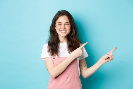 Image of cute teenage girl smiling, pointing fingers right at promo offer, demonstrating banner on copy space, standing in summer clothes with glamour make-up against blue background.