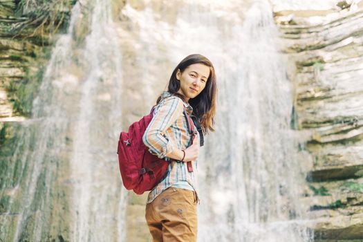 Explorer backpacker young woman standing on background of waterfall and looking at camera in summer outdoor.